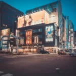 City street scene at dusk with brightly lit buildings, large advertising billboards, and cars at a crosswalk.
