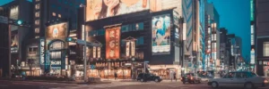 City street scene at dusk with brightly lit buildings, large advertising billboards, and cars at a crosswalk.