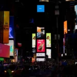 A nighttime scene at Times Square displaying brightly lit digital billboards and advertisements, with people and traffic below.