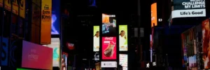 A nighttime scene at Times Square displaying brightly lit digital billboards and advertisements, with people and traffic below.