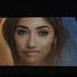 A close-up image of a woman's face displayed on a large electronic screen in a dark setting.