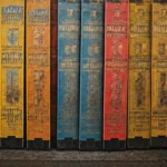 A row of worn, colorful hardcover books with faded spines lined up vertically on a shelf.