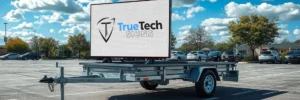 A trailer-mounted digital sign displaying the TrueTech Signs logo in a parking lot on a partly cloudy day.
